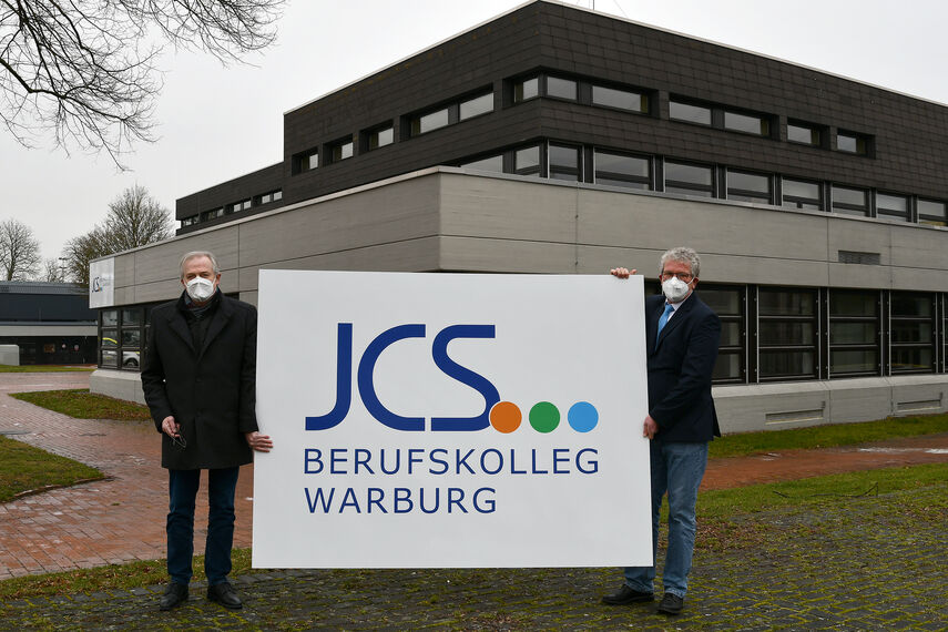 Kreisdirektor Klaus Schumacher und Schulleiter Matthias Gehle zeigen auf einem großen Schild das neue Logo des Johann-Conrad-Schlaun-Berufskollegs Warburg. Sie stehen mit dem Schild vor dem Berufskolleg in Warburg.