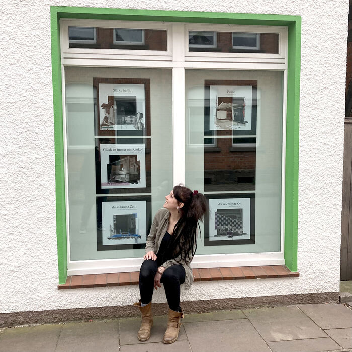 Eine junge Dame sitzt vor einem Fenster und schaut sich die Werke eines Künstlers an, die im Schaufenster dekoriert sind.
