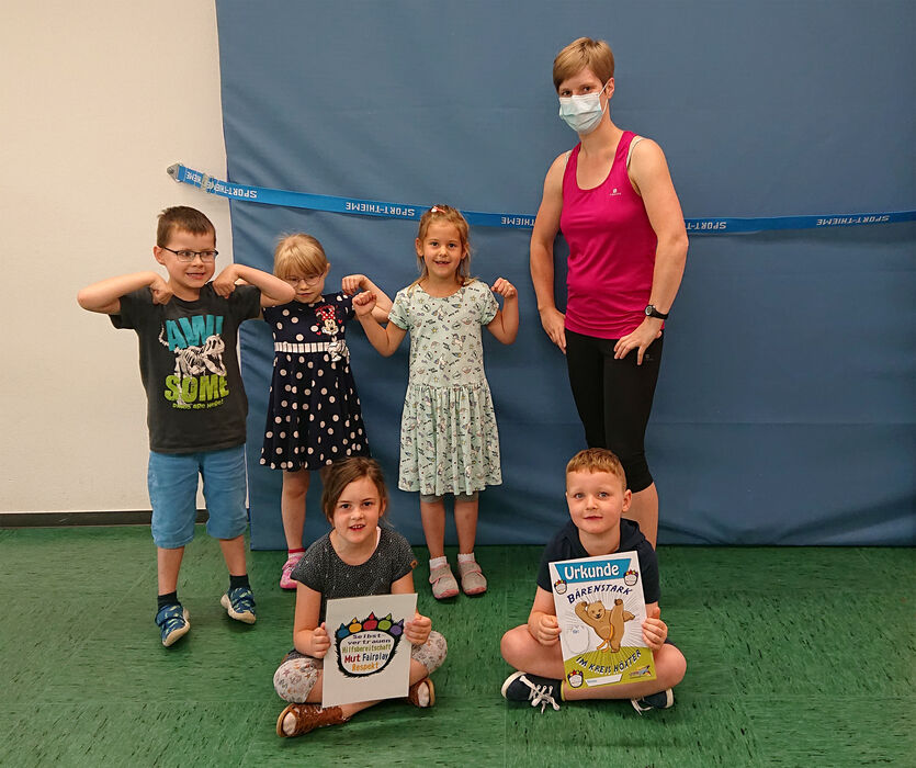 Die Kursleiterin Petra Koch steht mit 5 Kindern in einer Turnhalle. Die Kinder halten die Urkunde 'Bärenstark' in der Hand.
